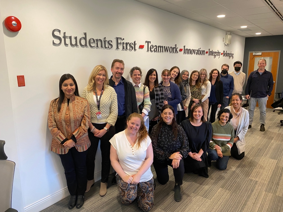 The 16 new faculty members stand in the board room underneath the college's values of Students First, Teamwork, Innovation, Integrity, and Belonging
